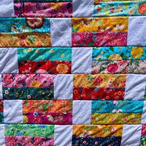 May include: A colorful patchwork quilt with a floral pattern. The quilt features a variety of fabrics in shades of pink, yellow, blue, and green, with white rectangular blocks separating the patterned sections. The quilt is a handmade item.