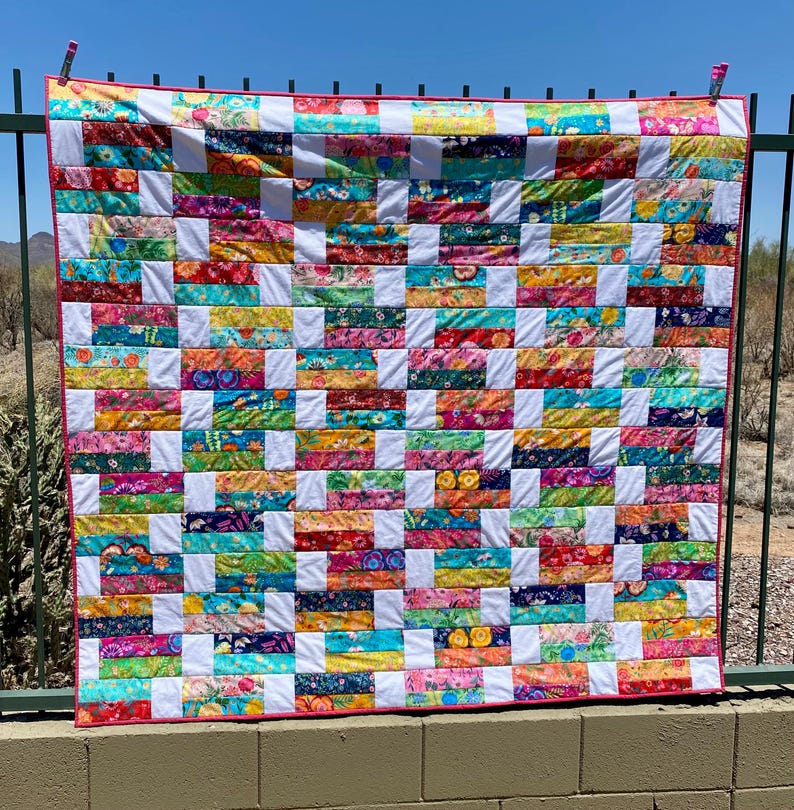 May include: A colorful handmade quilt with a patchwork design. The quilt features rectangular blocks of vibrant floral fabrics in shades of red, blue, yellow, and green, separated by white squares. The quilt is trimmed with a pink border and is hanging outside.