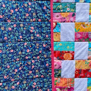 May include: A colorful quilt with a floral pattern. The left side features a dark blue fabric with pink flowers and green leaves. The right side has a patchwork design with various floral fabrics and white squares, separated by a pink border.