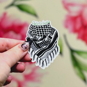 May include: A die-cut sticker featuring a black and white illustration of a keffiyeh scarf with a fringed edge. The word Palestine is written in green cursive text at the top of the sticker, accompanied by a small green heart symbol.