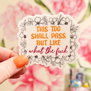 May include: A rectangular sticker with a scalloped edge featuring a black line art floral border. The text in orange reads THIS TOO SHALL PASS BUT LIKE and the text in dark pink reads what the fuck in a cursive script font.