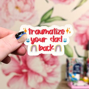 May include: A white die-cut sticker featuring the red text "traumatize your dad back" in a bold, rounded font. The design includes two orange four-pointed stars, two blue smiley faces, and two small rainbow icons at the bottom of the sticker.