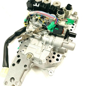 Puede incluir: Una unidad de control de transmisión automática plateada con múltiples conectores eléctricos y cables. La unidad tiene una cubierta de plástico negro y una carcasa de metal plateado.