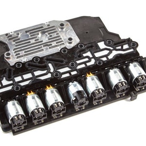 May include: A black automotive transmission valve body with six silver solenoids. The solenoids have gold-coloured electrical connectors.
