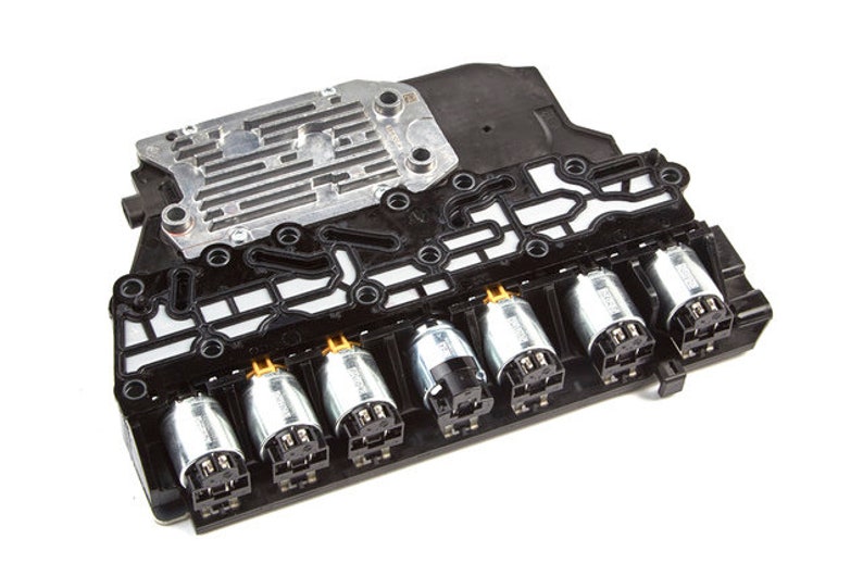 以下が含まれることがあります： 黒い自動車用トランスミッションバルブボディのクローズアップ。6つの銀色のソレノイドが取り付けられています。ソレノイドには金色の電気コネクターが付いています。