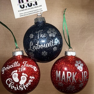 Puede incluir: Tres adornos navideños redondos con acabados de purpurina. Uno azul oscuro dice The Leibowitzs con copos de nieve. Dos rojos tienen texto blanco que dice Priscilla James 1st Christmas con huellas de bebé y MARK JR con copos de nieve.
