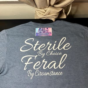 Könnte beinhalten: Ein heideblaues T-Shirt mit weißem Text, der Sterile By Choice Feral By Circumstance lautet. Eine kleine rechteckige Visitenkarte mit dem Text DCI Customize It befindet sich in der Nähe des Halsausschnitts des T-Shirts.