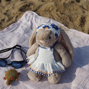 Può includere: Un coniglio di peluche che indossa un vestito bianco all'uncinetto con dettagli blu e arancioni. Il coniglio indossa un cappello all'uncinetto blu e bianco. Il coniglio è seduto su una coperta bianca con una piccola tartaruga all'uncinetto accanto a lui.