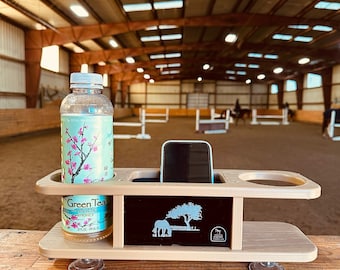 May include: A beige wooden phone and drink holder with suction cups on the bottom. The holder has a compartment for a phone, a cup holder, and a bottle holder. A bottle of green tea with ginseng and honey is in the bottle holder. A black phone is in the phone holder. The holder is sitting on a wooden surface. The background is a blurry image of an indoor riding arena.