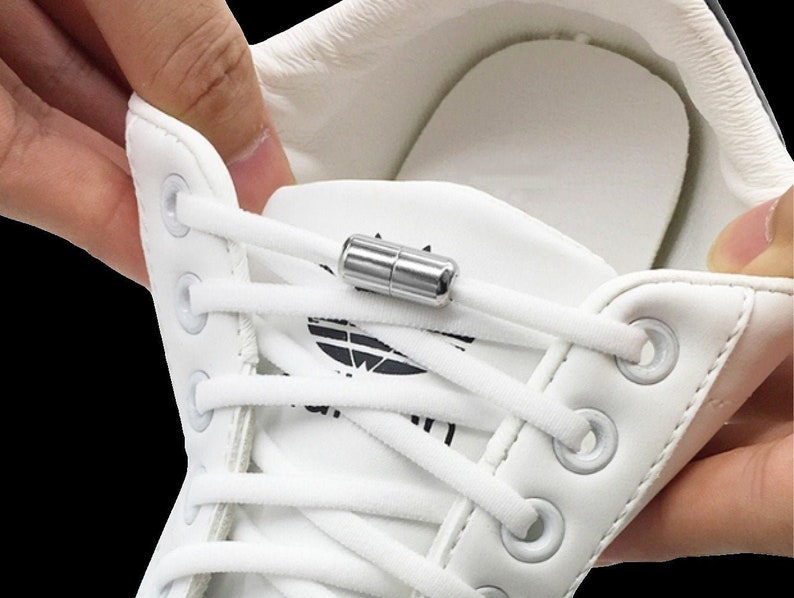 Puede incluir: Zapatillas blancas con cordones blancos y un cierre magn&eacute;tico plateado. Los cordones est&aacute;n enhebrados a trav&eacute;s de los ojales del zapato.