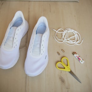 Puede incluir: Un par de zapatillas blancas con un cord&oacute;n blanco, dos ojales met&aacute;licos plateados, un par de tijeras amarillas y un encendedor colorido sobre una superficie de madera.