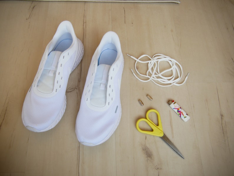 Puede incluir: Un par de zapatillas blancas con un cord&oacute;n blanco, dos ojales met&aacute;licos plateados, un par de tijeras amarillas y un encendedor colorido sobre una superficie de madera.