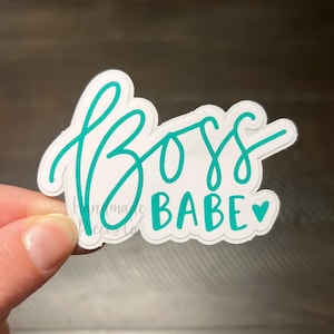 Puede incluir: Una pegatina blanca con las palabras "Boss Babe" en escritura turquesa. La palabra "Boss" está en una fuente grande y fluida, y "Babe" en una fuente más pequeña con un corazón turquesa. La pegatina está sujeta por una mano.