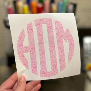 Puede incluir: Una calcoman&iacute;a de monograma rosa con fondo blanco. El monograma es un c&iacute;rculo con un borde festoneado y un patr&oacute;n de lazos rosas en el interior. Las letras "MOM" est&aacute;n recortadas del c&iacute;rculo.