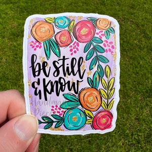 Puede incluir: Una pegatina rectangular con un diseño floral y las palabras "be still & know". La pegatina presenta flores naranjas, rosas y turquesas con hojas verdes y detalles dorados. El fondo es de color lila claro.