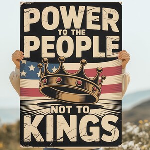 May include: A rectangular sign with the text "POWER TO THE PEOPLE NOT TO KINGS" in a distressed font. The sign features a crown over an American flag design. The sign is held by a person.