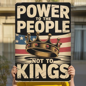 May include: A rectangular sign with the text "POWER TO THE PEOPLE NOT TO KINGS" in a distressed font. The sign features an American flag design and a gold crown illustration. The sign is held by a person wearing a yellow shirt.