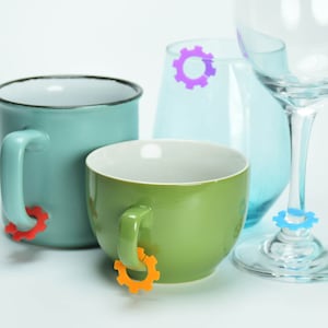 May include: Three mugs with colorful gear-shaped drink markers. The mugs are blue, green, and clear. The markers are red, orange, and purple.