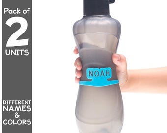 May include: A gray water bottle with a black cap and a blue band with the name "NOAH" printed on it. The bottle is held by a hand. Text on the left side says "Pack of 2 UNITS" and "DIFFERENT NAMES & COLORS".