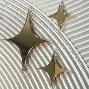 May include: Three gold star-shaped decorative objects with pointed edges are arranged on a textured white surface. The stars have a reflective, metallic finish, and the background has a ridged pattern.