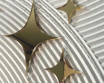 May include: Three gold star-shaped decorative objects with pointed edges are arranged on a textured white surface. The stars have a reflective, metallic finish, and the background has a ridged pattern.