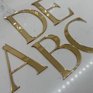 May include: Gold-colored, reflective acrylic letters A, B, C, D, and E are arranged on a white surface. The letters are in a serif font and appear to be cut from a sheet of gold-toned material. The letters are slightly angled.