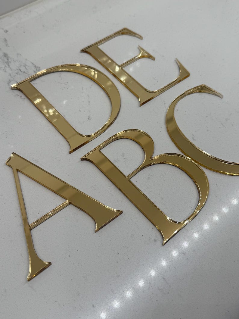 May include: Gold-colored, reflective acrylic letters A, B, C, D, and E are arranged on a white surface. The letters are in a serif font and appear to be cut from a sheet of gold-toned material. The letters are slightly angled.