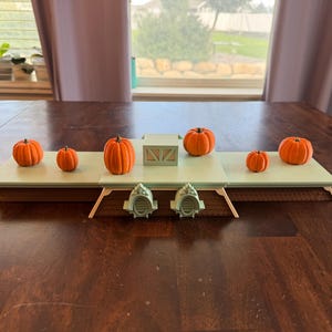 May include: A tiered display stand featuring a light green top surface and a brown base with two small arched architectural details. Six small orange pumpkins are arranged across the flat platforms of this tiered structure.