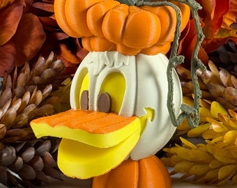 May include: A white pumpkin with a carved pumpkin face, a yellow beak, and orange feet. The pumpkin is topped with an orange pumpkin with a green stem and vine.