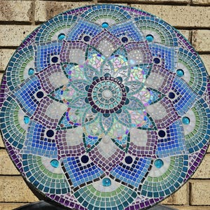 May include: A circular mosaic wall art featuring a mandala design with a flower in the center. The mandala is made of blue, green, purple, and white tiles. The flower in the center is iridescent.