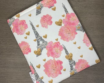 May include: A white fabric book sleeve featuring a pink and gold Eiffel Tower and floral design. The sleeve has the text "BookSaks" printed on the bottom left corner.