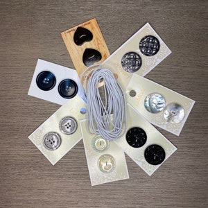 May include: Assortment of buttons in various colors, shapes, and sizes, including black, white, and silver. The buttons are displayed on cards with the word "Lamode" printed on them. There is also a spool of white elastic.