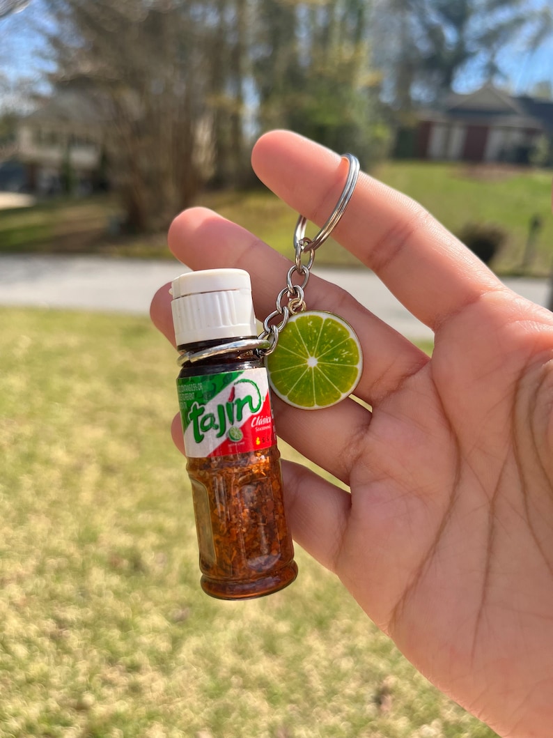 K&ouml;nnte beinhalten: Ein Schl&uuml;sselanh&auml;nger mit einer Miniaturflasche Taj&iacute;n-Gew&uuml;rz und einem Limetten-Anh&auml;nger. Die Flasche ist klar und zeigt das rote, gr&uuml;ne und wei&szlig;e Taj&iacute;n-Etikett. Der Limetten-Anh&auml;nger ist gr&uuml;n und gelb.