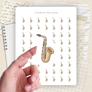 May include: A white sticker sheet featuring a repeating pattern of small saxophone illustrations and the text "L'armoire des profs". A larger saxophone illustration is centered. The image is held by a hand.