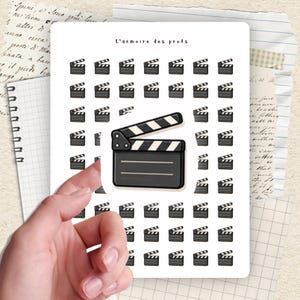 Peut inclure: Une planche d'autocollants avec un motif répété de cliquets de film. Le cliquet central est ouvert, avec des rayures noires et blanches. Le texte "L'armoire des profs" est en haut.