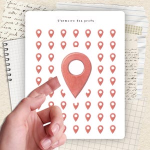 Puede incluir: Una tarjeta blanca con el texto "L'armoire des profs" y una cuadrícula de iconos de ubicación rojos. Una mano sostiene la tarjeta. El fondo incluye papel de cuaderno y papel vintage.