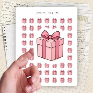 Peut inclure: Une feuille d'autocollants avec des boîtes cadeaux roses avec des nœuds. Le texte "L'armoire des profs" est en haut. Les autocollants sont sur fond blanc, une main tient la feuille.