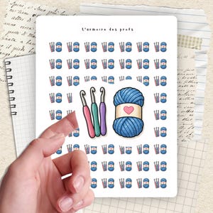 Peut inclure: Une planche d'autocollants avec des illustrations de pelotes de laine bleues avec des cœurs roses, des crochets et des aiguilles à tricoter. Le texte "L'armoire des profs" est en haut. La feuille est tenue par une main.