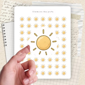 Peut inclure: Une feuille blanche avec un motif répété d'illustrations de soleil jaune. Le texte "L'armoire des profs" est en haut. Une main est partiellement visible, tenant la feuille. Le fond comprend du papier de cahier et des notes manuscrites.