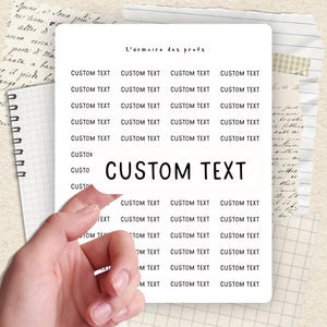 Peut inclure: Une feuille d'autocollants blanche avec le texte "CUSTOM TEXT" en gras. La feuille est tenue par une main et entourée d'autres textes et de papier de carnet. Les mots "L'armoire des profs" sont en haut.