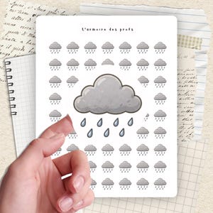 Könnte beinhalten: Ein Bogen mit Aufklebern mit grauen Wolken- und Regentropfen-Designs. Der Text "L'armoire des profs" steht oben. Die Aufkleber werden von einer Hand gehalten, mit einem Notizbuch und anderen Papieren im Hintergrund.