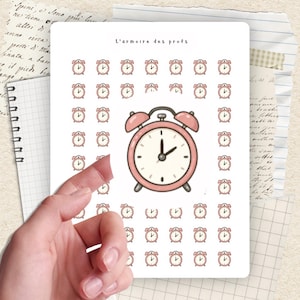 Peut inclure: Une planche d'autocollants avec une grande horloge rose et de nombreuses illustrations de petits réveils. Le texte "L'armoire des profs" est en haut. La feuille est tenue par une main.