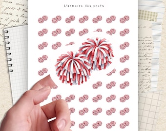 May include: A white sticker sheet with watercolor illustrations of red and white pom-poms. The text "L'armoire des profs" is at the top. The sticker sheet is held by a hand, with a notebook and paper in the background.
