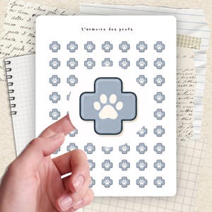 Puede incluir: Una hoja de pegatinas con temática veterinaria. Las pegatinas presentan una cruz azul claro con una huella de pata en el centro, rodeada de iconos de huellas de pata más pequeños. El texto "L'armoire des profs" está en la parte superior.