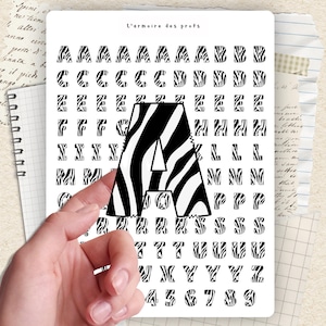 Peut inclure: Une planche d'autocollants blanche avec le texte L'armoire des profs en haut, comprenant plusieurs rangées de lettres de A à Z et de chiffres de 0 à 9 à motif zébré noir et blanc, avec une grande lettre A zébrée au centre de la feuille.