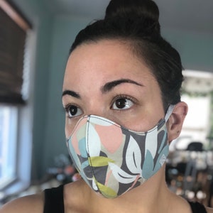 May include: A person wearing a face mask with a floral pattern in shades of gray, blue, green, and pink. The mask has a white background and is made of fabric.