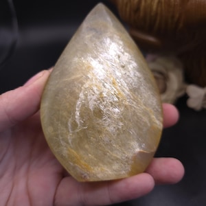 May include: A teardrop-shaped, translucent, light yellow crystal with a shimmery, iridescent finish. The crystal has a smooth, polished surface and is held in a hand.