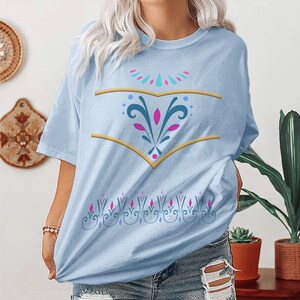 May include: A light blue t-shirt with a gold and blue design on the front. The design features a stylised floral pattern with pink accents.