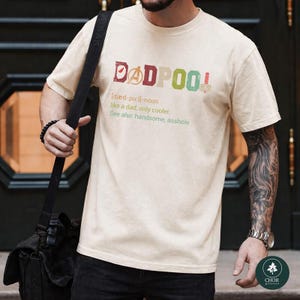May include: Cream-colored t-shirt with the text "DADPOOL" in red, green, and pink. Below the word is a definition: "like a dad, only cooler. See also: handsome, asshole."