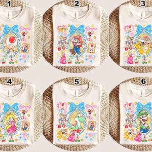 May include: Six off-white t-shirts with different cartoon character designs. Each shirt features a character in an oval frame with a blue bow, surrounded by floral and decorative elements. The characters include Toad, Mario, Bowser, Peach, Yoshi, and Luigi.